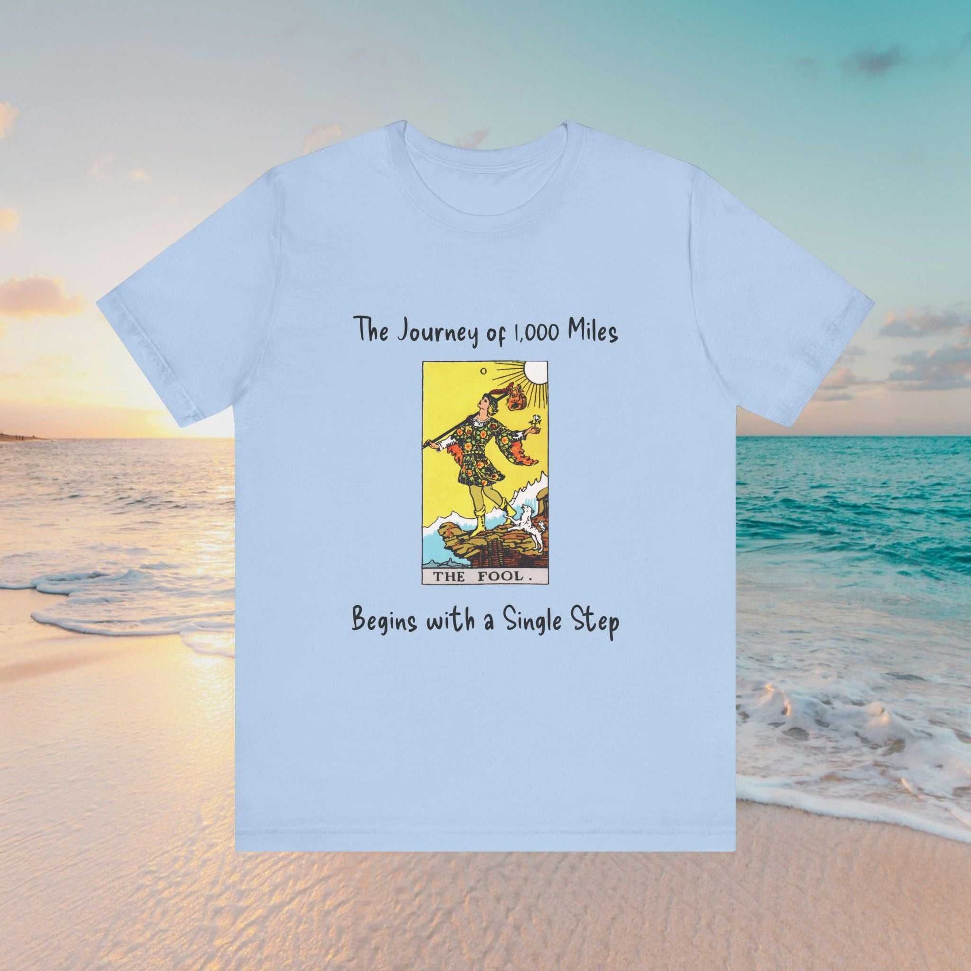 The Fool tarot graphic and text of 'The Journey of 1,000 Miles Begins with a Single Step" on a light blue tee
